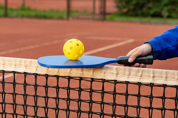 Was ist Pickleball erklärt – Pickleball Paddle und Ball auf dem Spielfeld Was ist Pickleball? Einfach erklärt. Gelber Lochball liegt auf blauem Paddle über dem Netz auf einem Spielfeld.