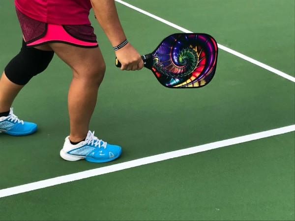 Pickleball Hamburg: Spieler bereitet Aufschlag auf grünem Platz vor, mit buntem Schläger und blauen Sportschuhen.